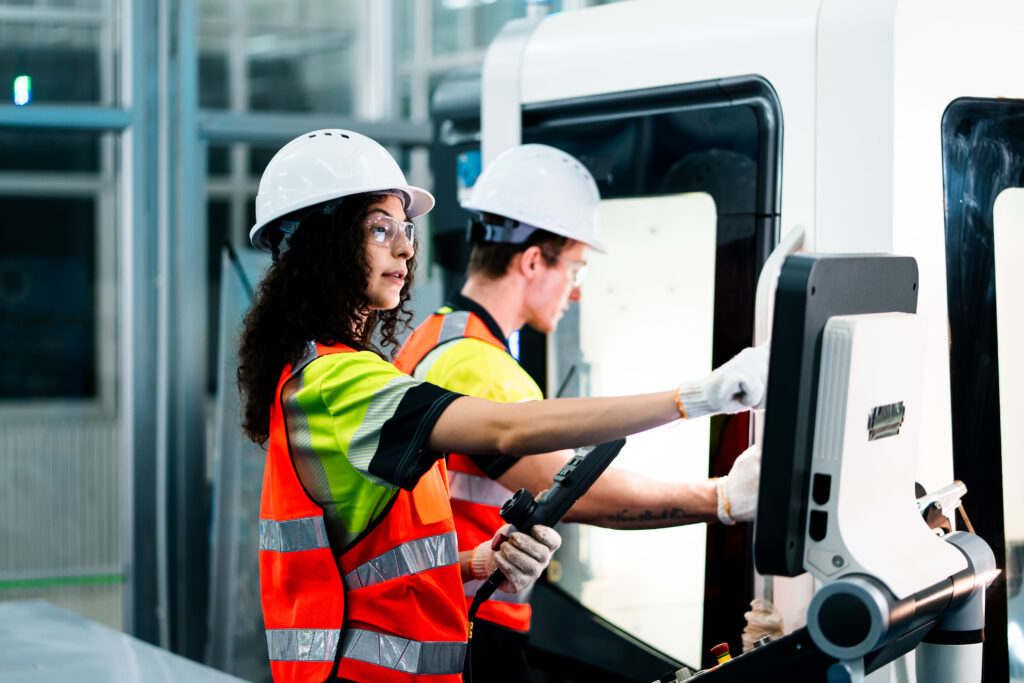 Two workers in orange vests are working on a machine. One of them is pointing at a screen